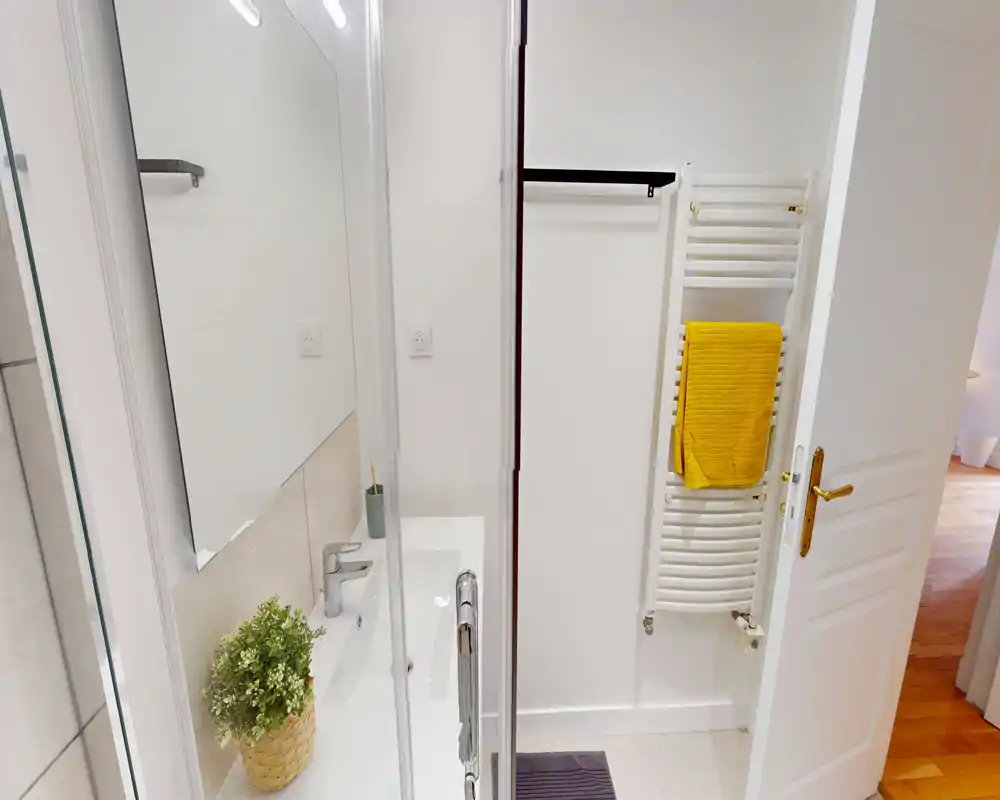 Compact, well-lit bathroom with a sink, mirror and a heated towel rack with a yellow towel. Clean white finishes and a small decorative plant add a fresh touch.