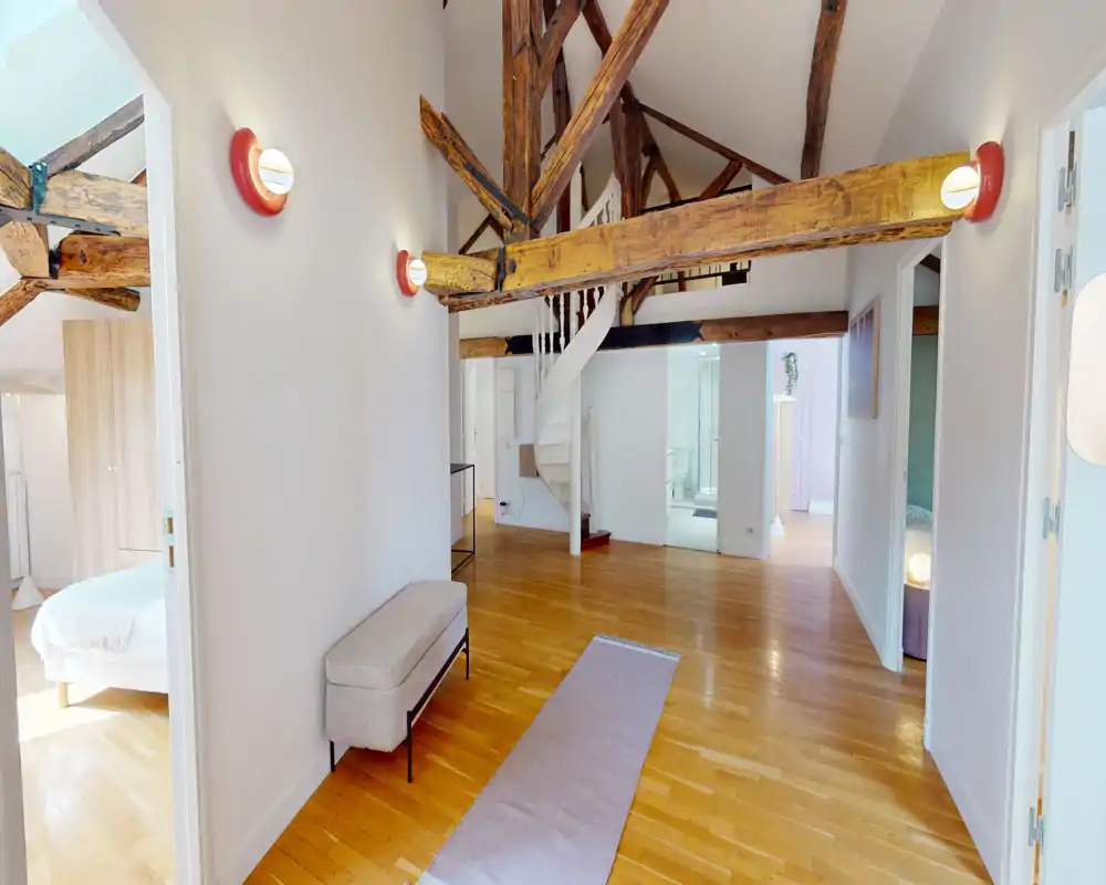 Bright, open living area with exposed wooden beams, warm hardwood floors, a small bench and decorative rug; modern and airy loft-style space.