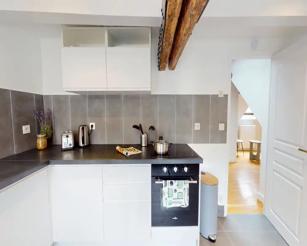 Modern compact kitchen with dark countertop, built-in oven, kettle and toaster; neutral tiles and exposed wooden beams create a clean, stylish space.