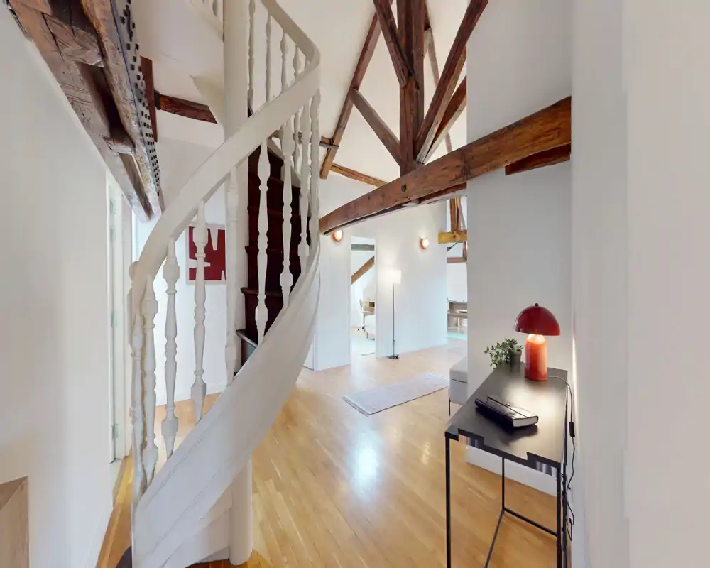 Bright, airy living area with exposed wooden beams, a decorative spiral staircase and a small console table with lamp — ideal as an open landing or lounge space.