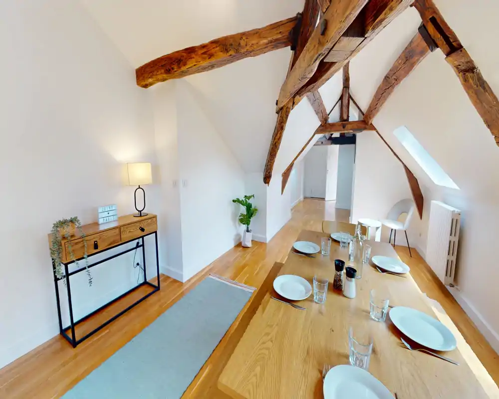 Stylish attic dining area with a long wooden table set for guests, warm hardwood floors and striking exposed wooden beams that give the space character.