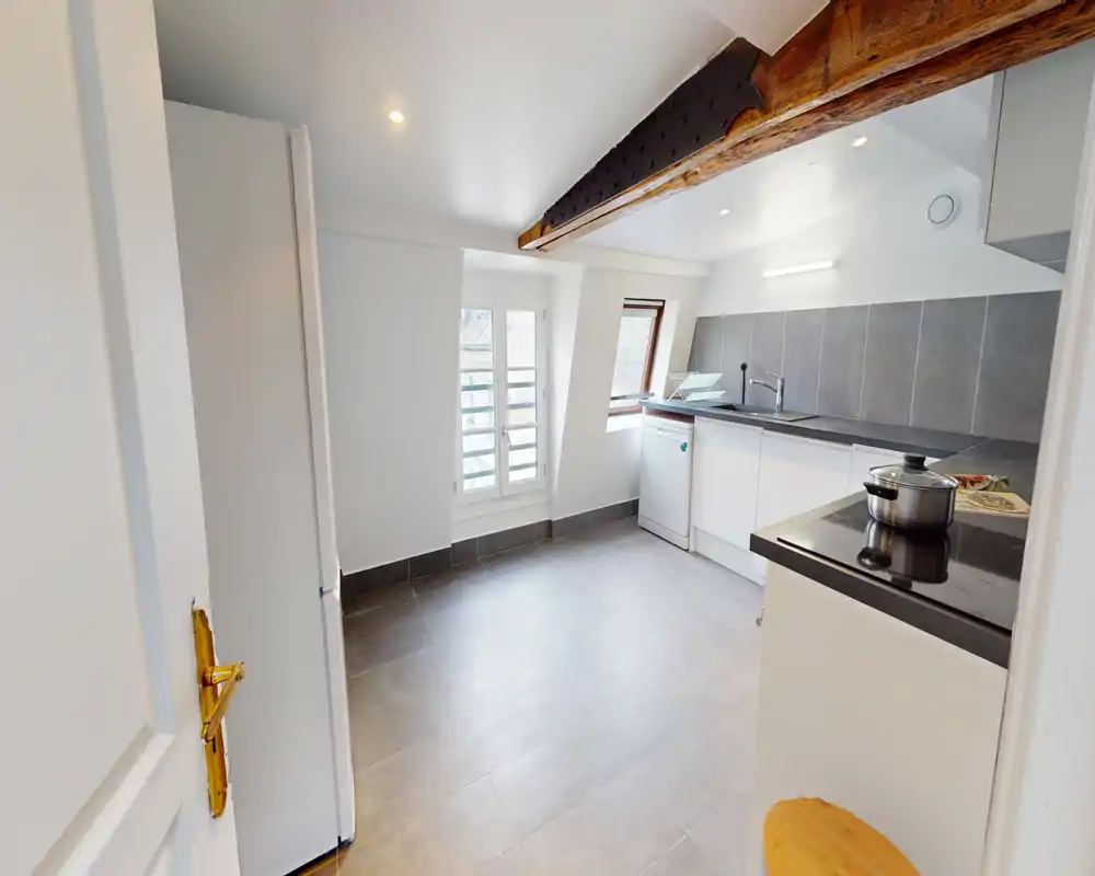 Compact, modern kitchen with white cabinets, dark countertop and tiled backsplash. Natural light from the window brightens the space and exposed wooden beam adds character.