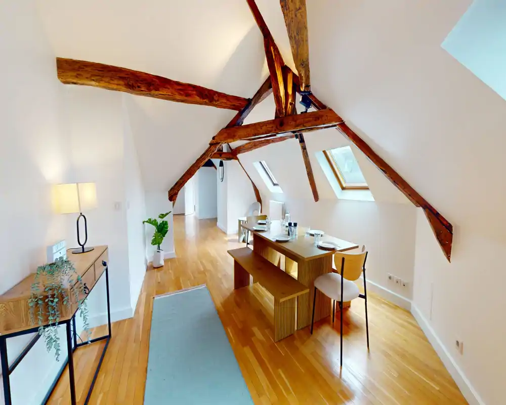 Bright attic dining area with exposed wooden beams, a wooden bench-style table set under skylights and warm parquet flooring — a cozy spot for meals.