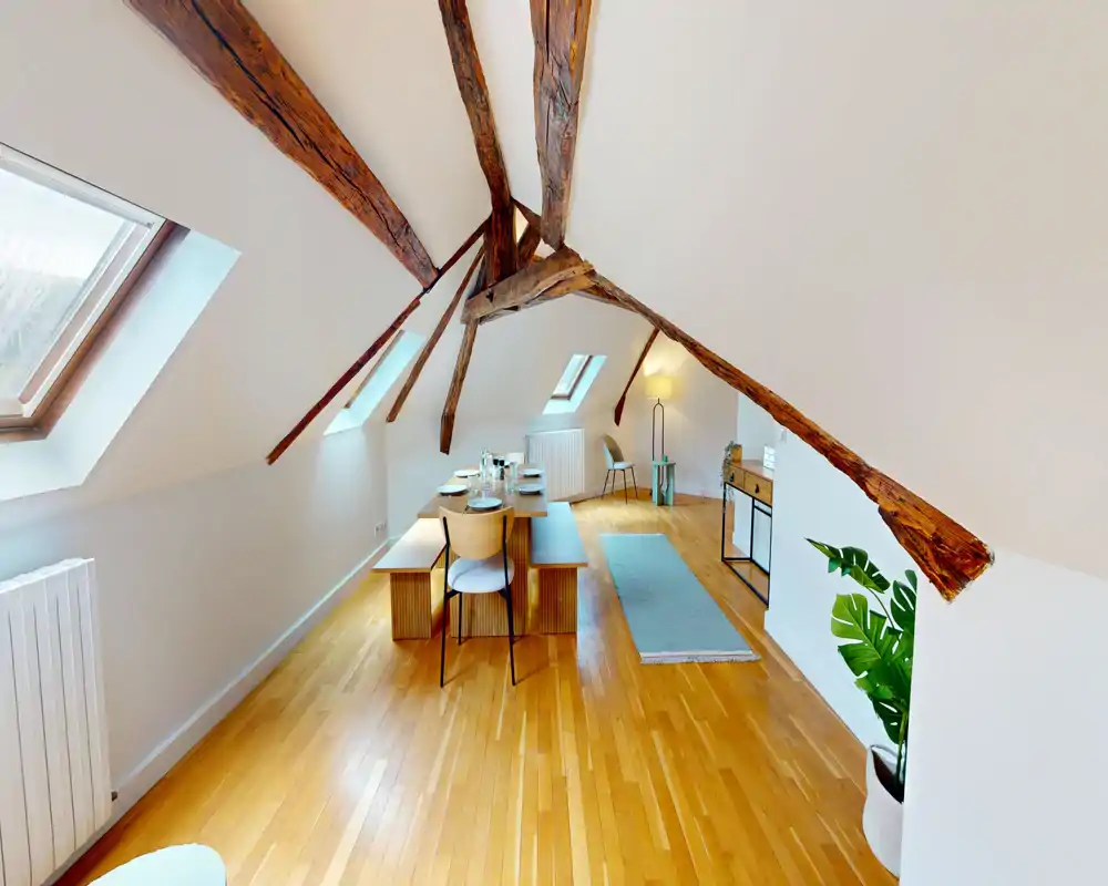 Attic-style dining area under exposed wooden beams with a long table, bench seating and skylights bringing abundant natural light—warm wood floors and tasteful decor create a welcoming dining space.