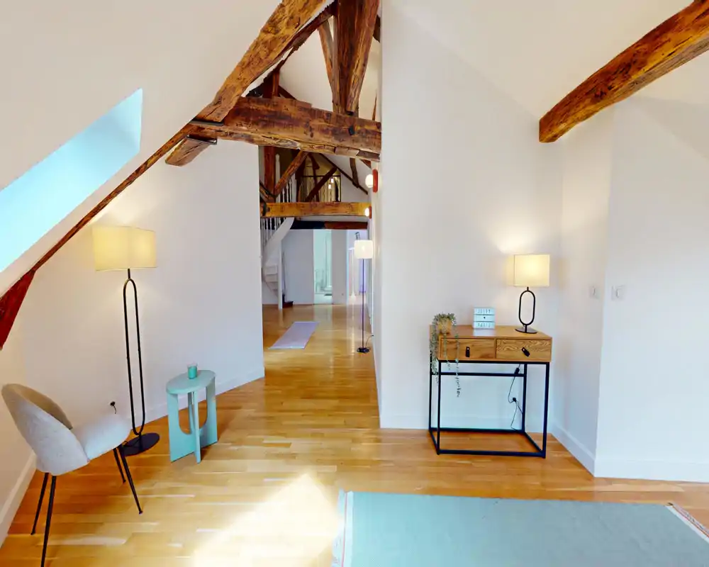 Bright, stylish living area with exposed wooden beams, warm hardwood floors, a cozy chair and soft lighting — a welcoming open-plan space.