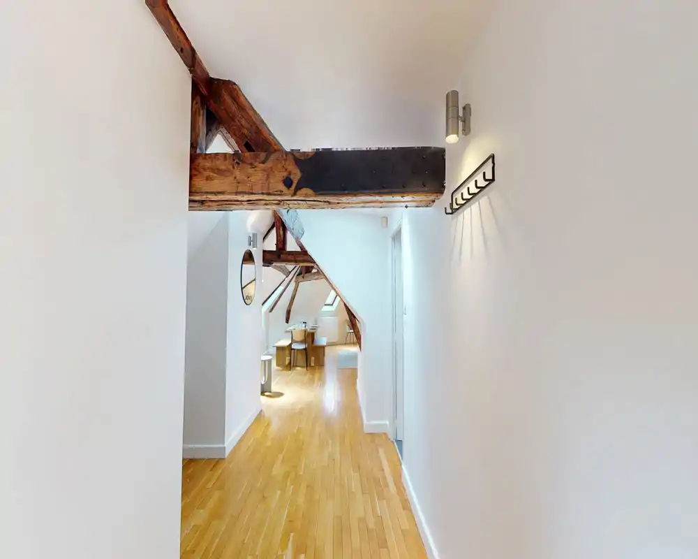 Bright, airy attic living area with exposed wooden beams, light hardwood floors and a small dining/work corner; modern minimalist decor and good natural light.