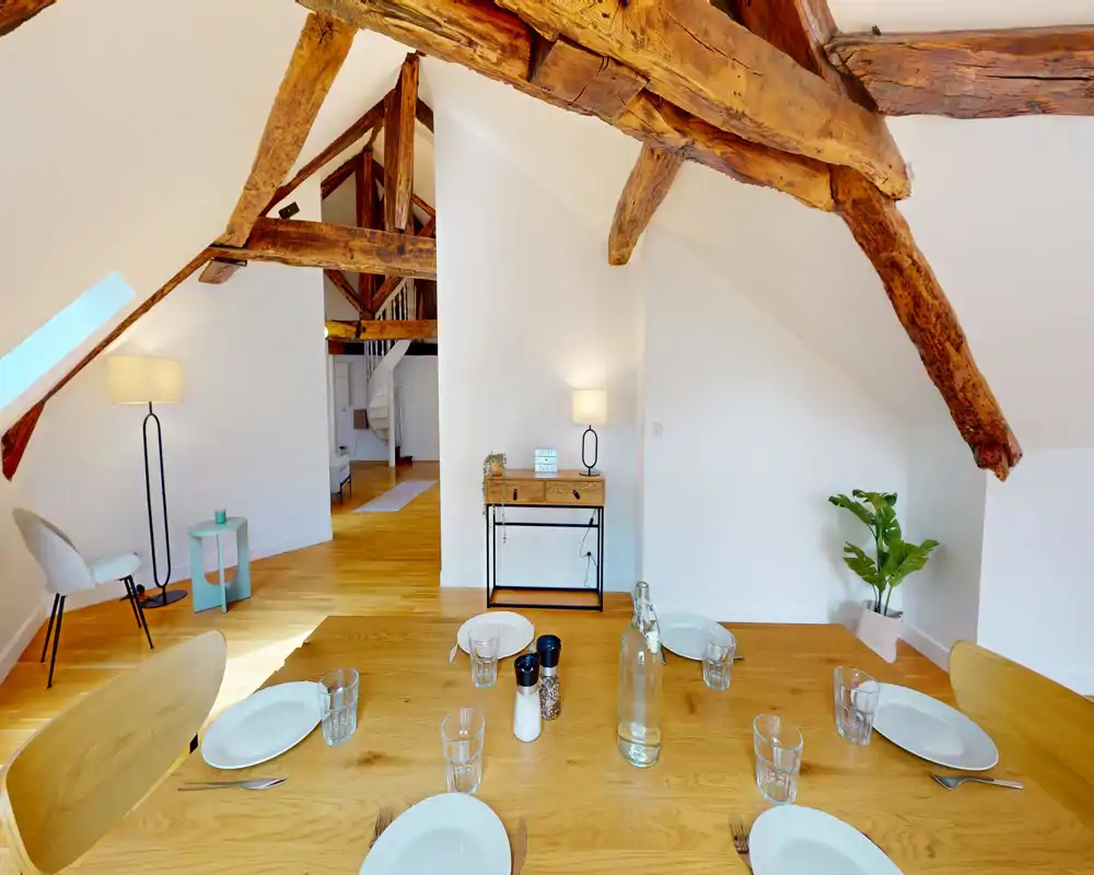 Bright attic dining area with a wooden table set for four, exposed rustic beams and contemporary chairs — warm, airy and inviting.
