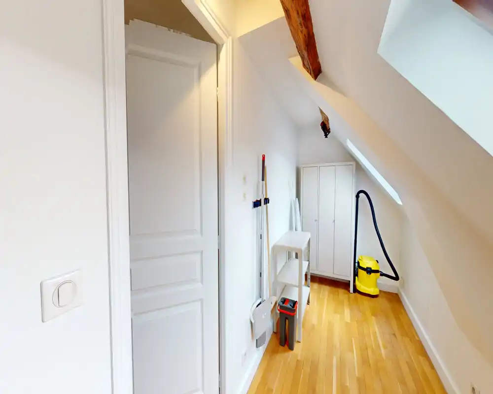 Narrow attic corridor used as a storage/entrance area with wooden floor, sloped ceiling and a small wardrobe; includes cleaning tools and a vacuum cleaner.