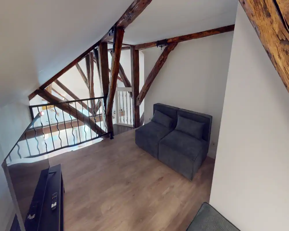 A cozy mezzanine living area with exposed wooden beams, a compact two-seat sofa and light wood flooring; ideal as a quiet lounge or reading nook.