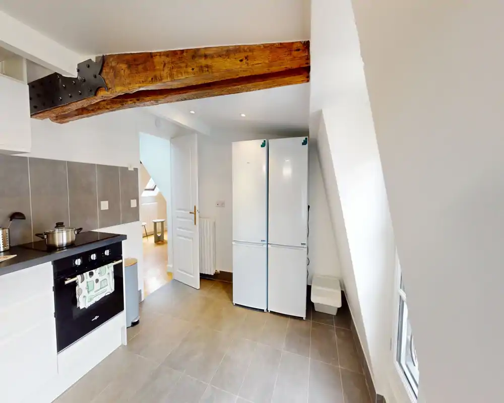 Bright, modern kitchen with built-in oven, countertop workspace and a tall double fridge under an exposed wooden beam. Clean tiled floor and neutral finishes create a practical cooking area.