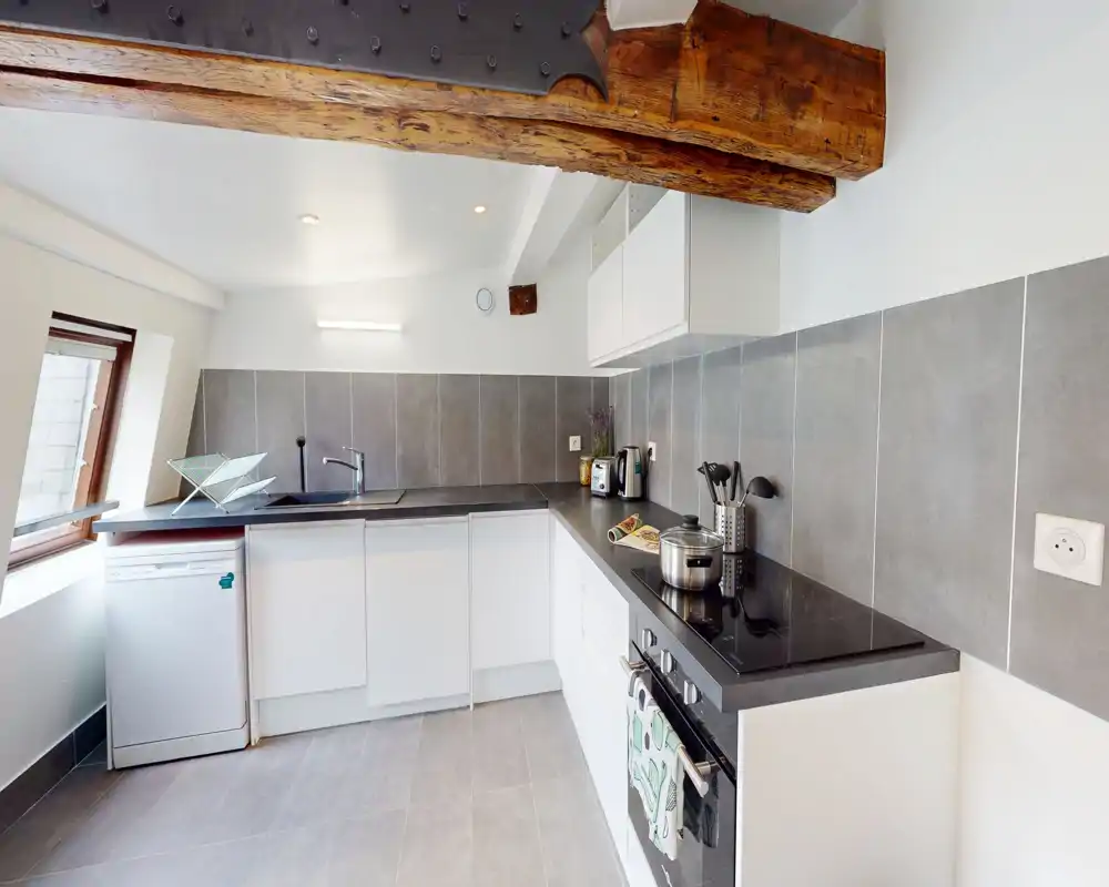 Modern compact kitchen with white cabinetry, dark countertops and integrated appliances; natural light from a side window and exposed wooden beam add character.