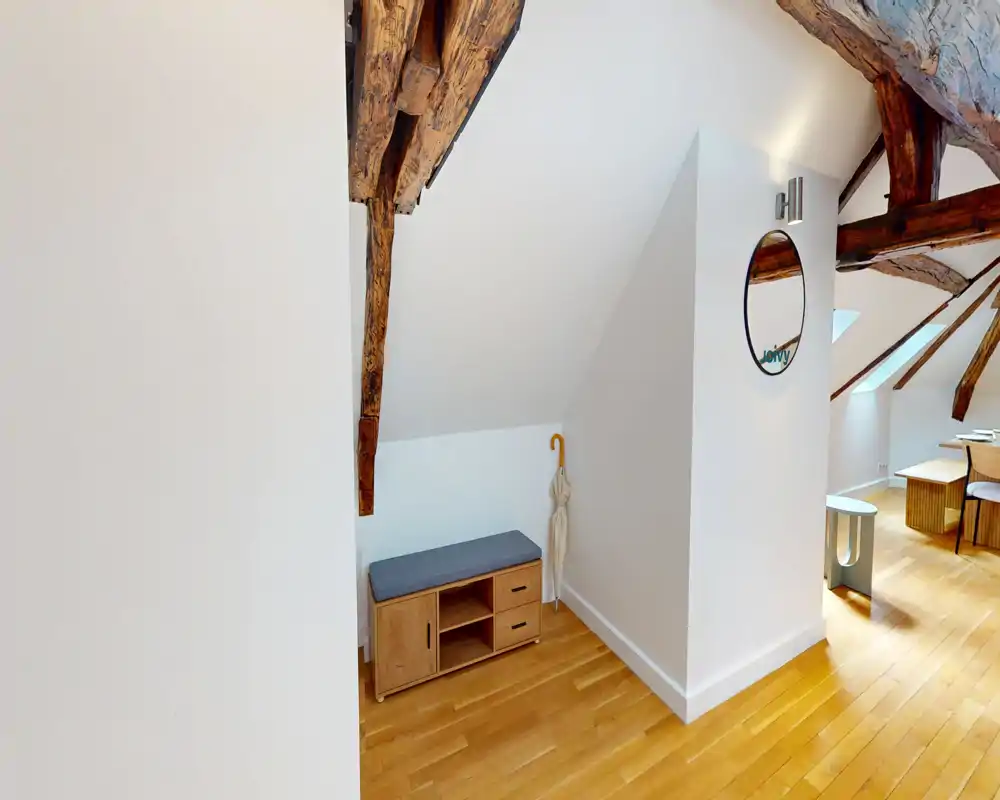 Compact entrance/landing under exposed wooden beams with a small bench, umbrella stand and mirror; bright parquet flooring and a neat, welcoming feel.
