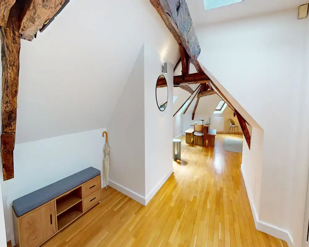 Bright attic living space with exposed wooden beams, light hardwood floors and minimalist furnishings; comfortable seating and a small dining area make this a pleasant open-plan lounge.