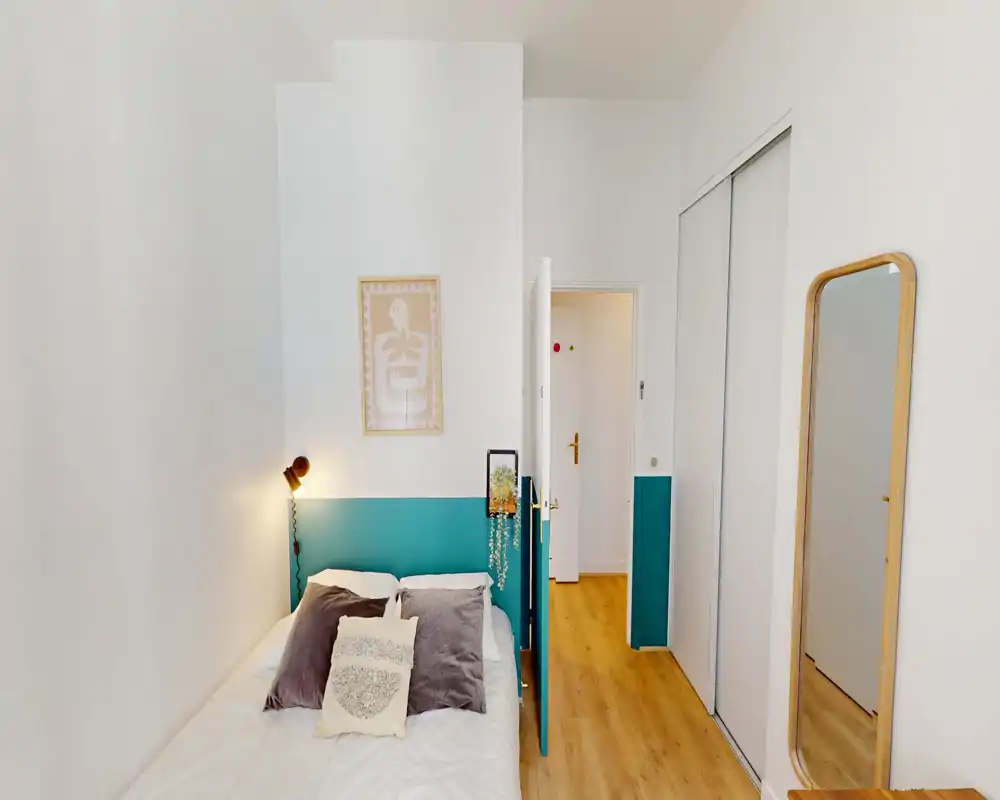 Camera da letto compatta e luminosa con testiera dipinta di turchese, letto singolo con biancheria neutra, cuscini decorativi e lampada da parete. Stile pulito e minimal con specchio a figura intera per ampliare lo spazio.