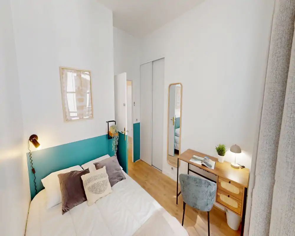 Camera da letto luminosa e ben arredata con un comodo letto matrimoniale, cuscini decorativi e una lampada da comodino accogliente. Una scrivania in legno e uno specchio creano un angolo lavoro funzionale vicino alla finestra.
