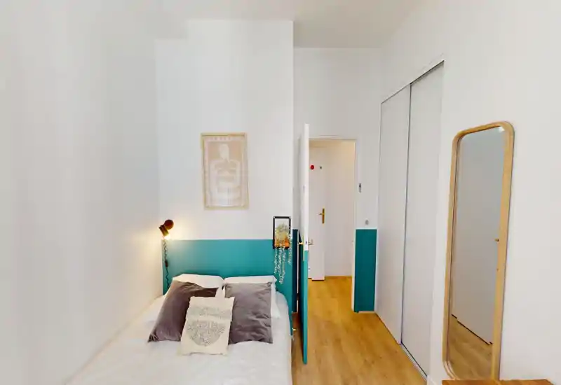 Camera da letto compatta e luminosa con testiera dipinta di turchese, letto singolo con biancheria neutra, cuscini decorativi e lampada da parete. Stile pulito e minimal con specchio a figura intera per ampliare lo spazio.
