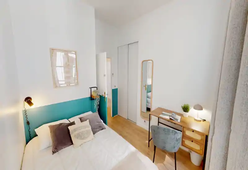 Camera da letto luminosa e ben arredata con un comodo letto matrimoniale, cuscini decorativi e una lampada da comodino accogliente. Una scrivania in legno e uno specchio creano un angolo lavoro funzionale vicino alla finestra.