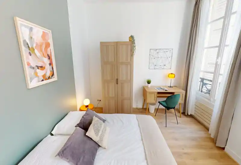 Bright, stylish bedroom with a large bed, bedside lamps, wooden wardrobe and a small desk by the window — airy natural light and soft textiles create a welcoming space.