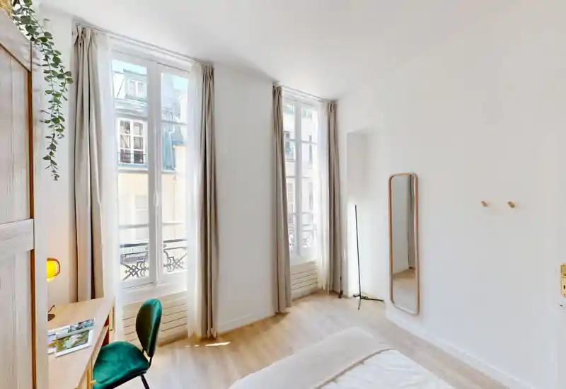 Bright, minimalist bedroom with tall windows, light wood floors and a partially visible bed in the foreground; a small desk, green chair and full-length mirror complete the airy space.