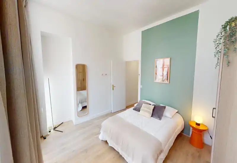 Bright, stylish bedroom with a double bed against a green accent wall, cozy textiles and a bedside lamp creating a warm atmosphere. Large window provides abundant natural light and pale wooden floors add a modern touch.