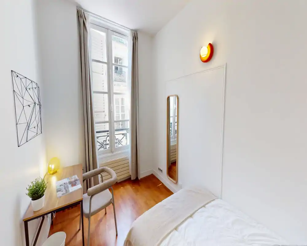 Camera da letto luminosa e accogliente con letto rifatto, pavimento in legno e scrivania accanto a una finestra alta che lascia entrare molta luce naturale. Arredo minimal, specchio a figura intera e tonalità morbide ideali per un annuncio di affitto.