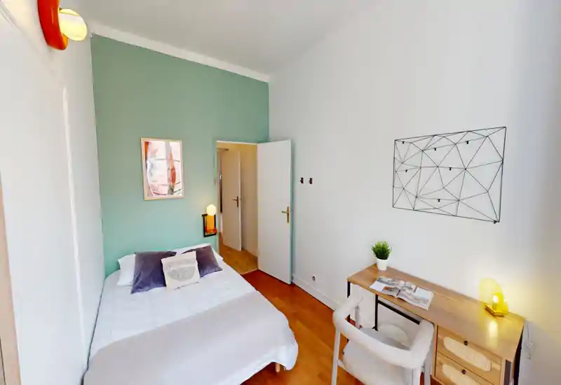 Camera da letto compatta e luminosa con letto ben rifatto contro una parete verde salvia, lampada da comodino e un piccolo angolo studio con scrivania e sedia — perfetta per viaggiatori singoli o affitti accoglienti.