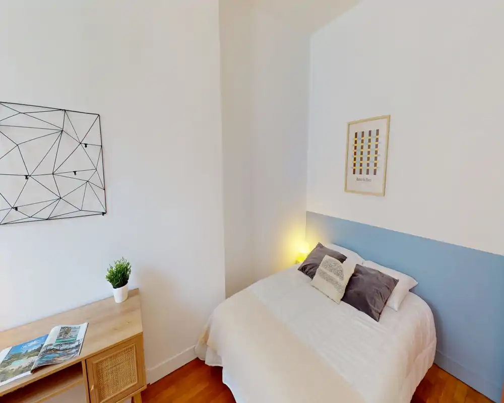 Camera da letto luminosa e accogliente con letto matrimoniale ben rifatto, cuscini decorativi e lampada da comodino. Arredamento minimalista, pavimento in legno e piccolo tavolino laterale.