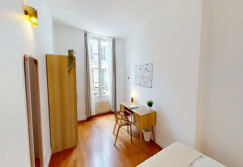 Camera da letto luminosa e compatta con pavimento in legno e una piccola scrivania vicino alla finestra. Armadio semplice, specchio a tutta altezza e luce calda creano uno spazio ordinato e funzionale, ideale per soggiorni brevi.