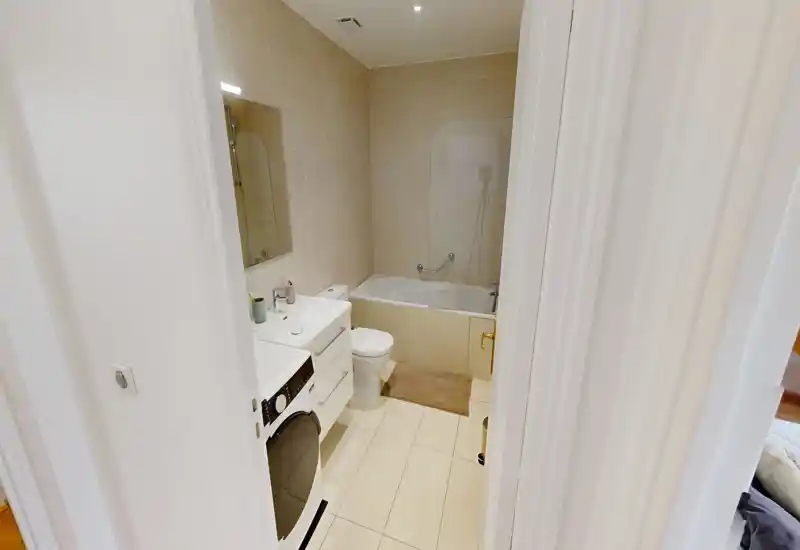Compact, well-lit bathroom with a bathtub, toilet, sink and a washing machine tucked under the counter. Neutral tiles and clean fittings create a neat, modern look.