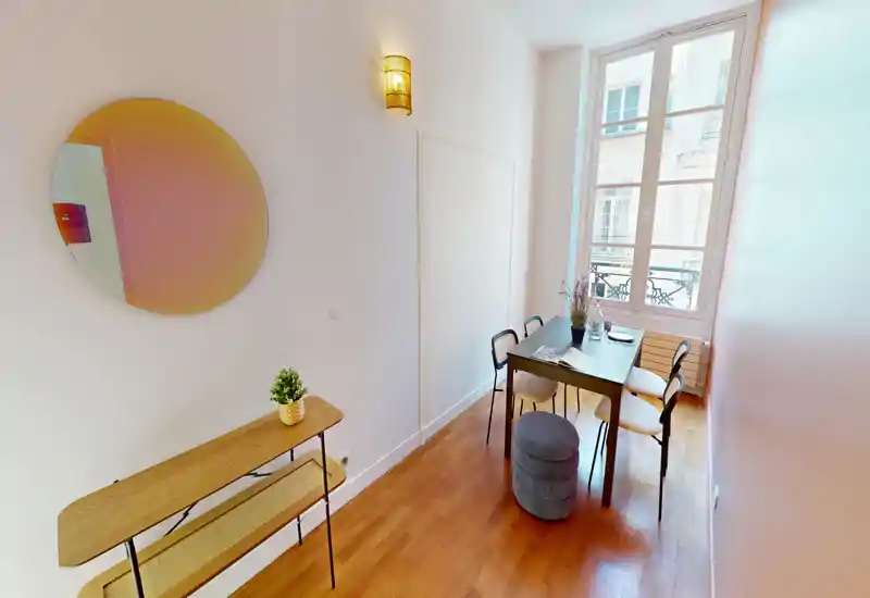 Compact dining area with a wooden table for four by a large window, warm hardwood floors and tasteful minimal decor — ideal for cozy meals and bright natural light.