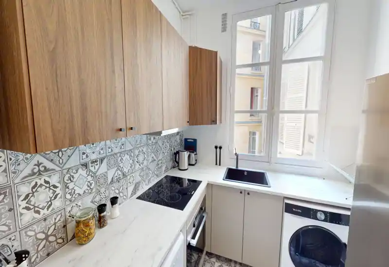 Cucina moderna e luminosa con paraschizzi a piastrelle decorative, pensili in legno, piano cottura a induzione, lavello sotto una grande finestra e lavatrice integrata — compatta e di stile per la vita in città.