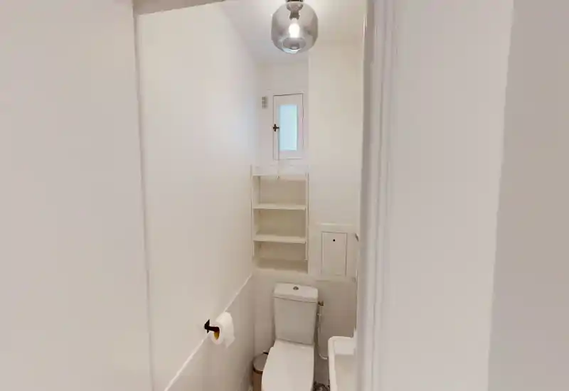Compact, all-white bathroom with a toilet, small sink and built-in shelving under a small window. Bright, minimal and freshly painted, suitable for single occupancy or a guest WC.