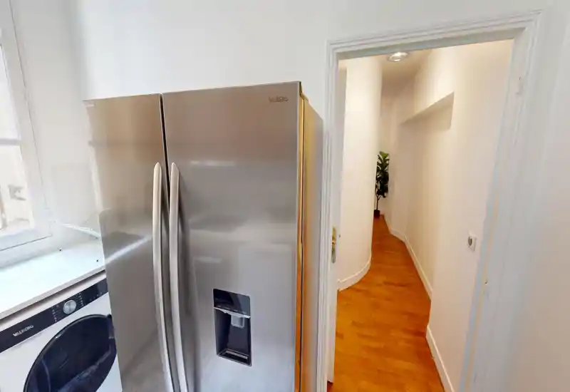 Compact kitchen/laundry corner with a stainless-steel side-by-side refrigerator and a front-loading washer next to a bright window; wooden floors and a corridor visible beyond.