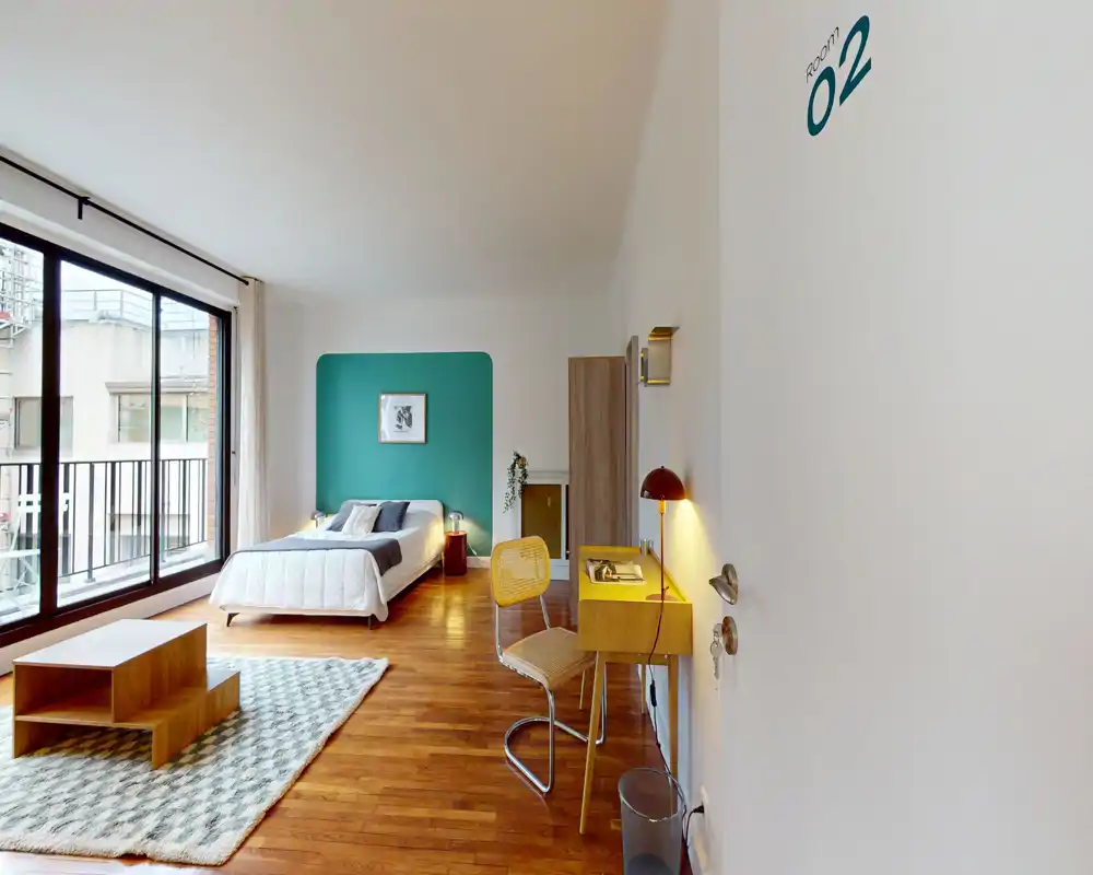 Bright, modern bedroom with a double bed against a teal accent wall, large floor-to-ceiling windows, wooden flooring and a small desk area — ideal for a comfortable city stay.