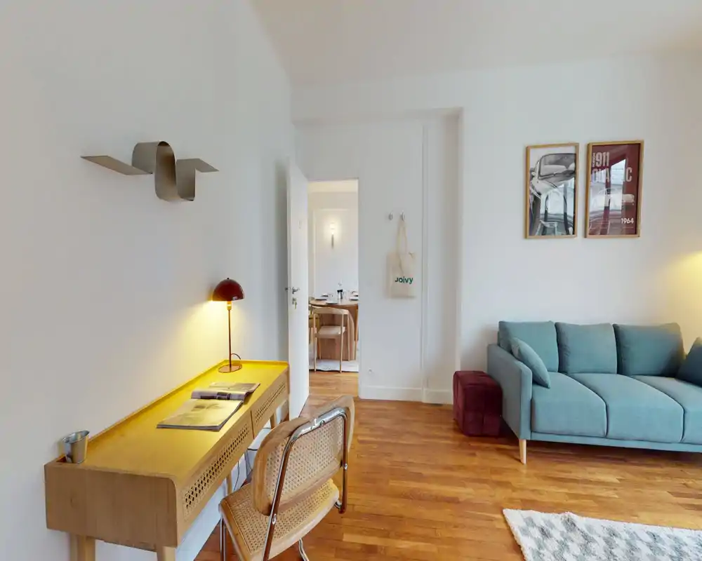 Bright, well-staged living room with a teal sofa, wooden flooring and a compact desk area — warm lighting and tasteful decor create a welcoming atmosphere.