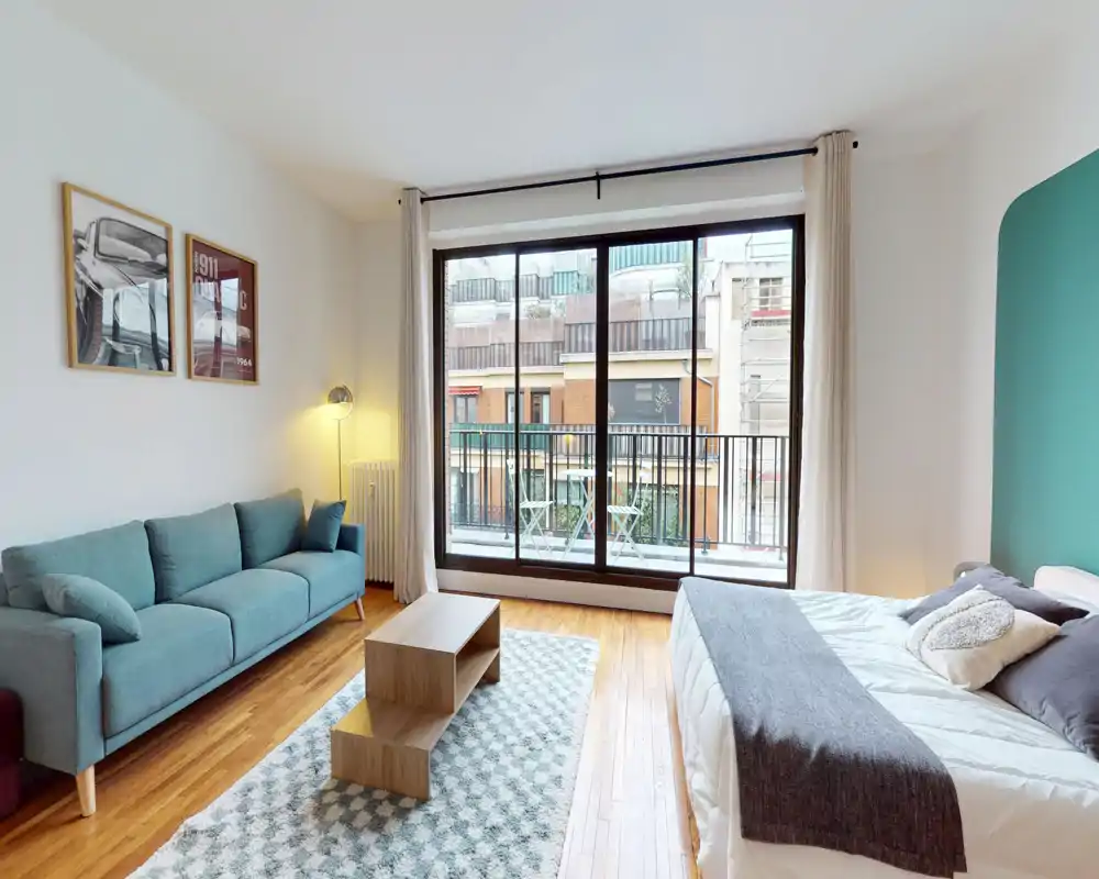 Bright, well-staged studio bedroom with a comfortable double bed, a teal sofa, wooden floors and large floor-to-ceiling windows opening onto a balcony.