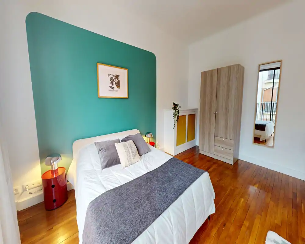 Bright, modern bedroom with a teal accent wall, double bed dressed in white linens and grey throws, bedside lamps and a wardrobe. Warm wooden floors and a full-length mirror complete the cozy, well-lit room.