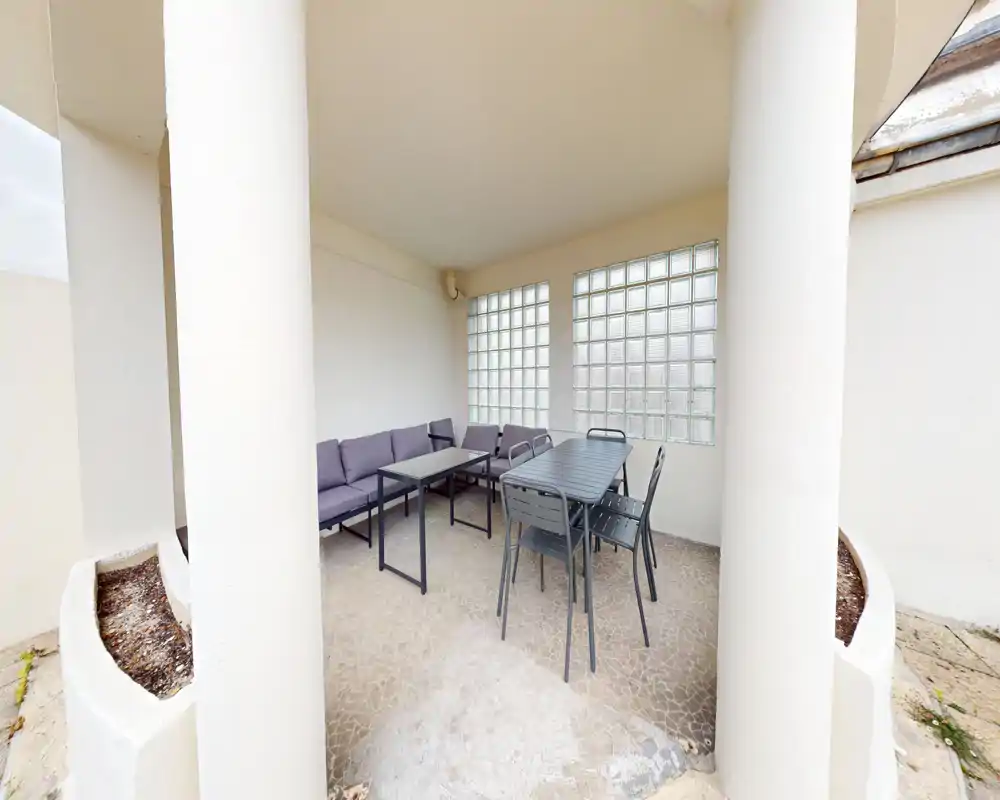 Espace extérieur couvert avec table à manger en métal, chaises et canapé d'angle rembourré sous un patio abrité — idéal pour dîner en plein air ou se détendre.