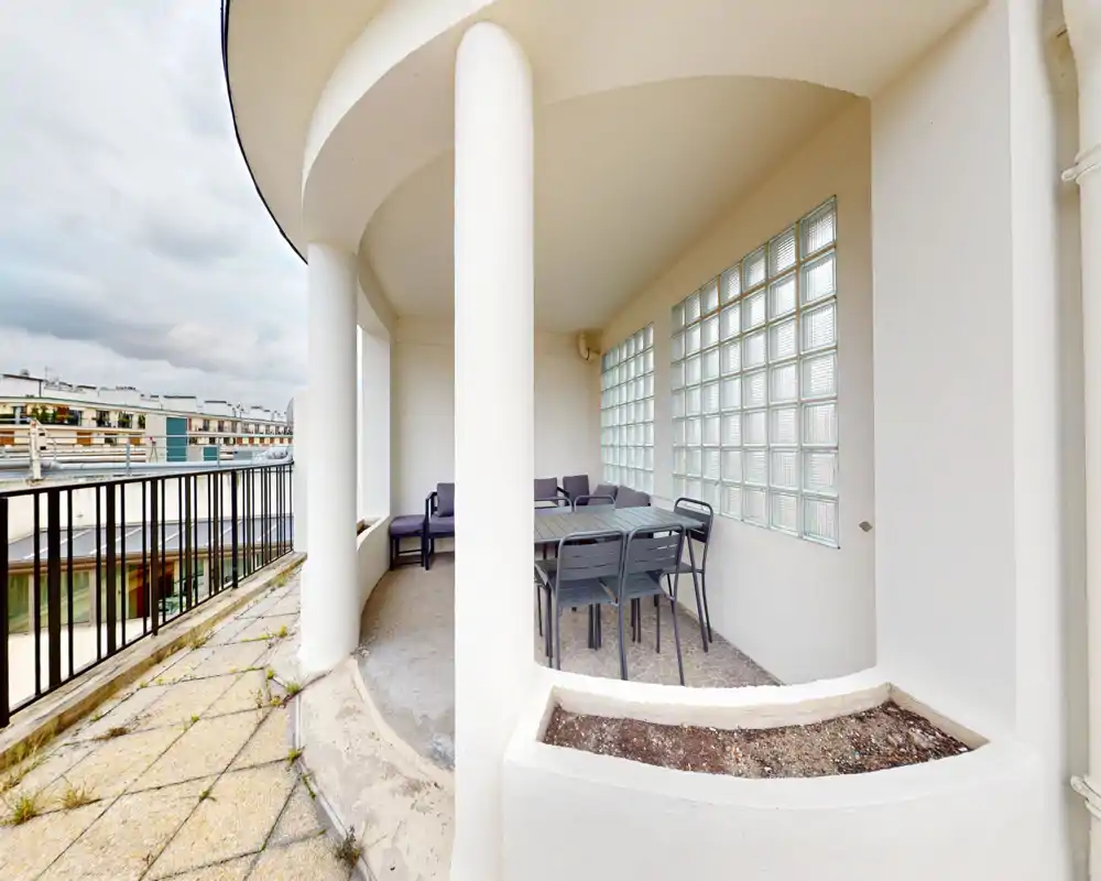 Terrasse couverte avec table et chaises et petit coin salon, abritée par des colonnes courbes et des vitres en verre brique — un espace extérieur privé adapté aux repas ou à la détente.