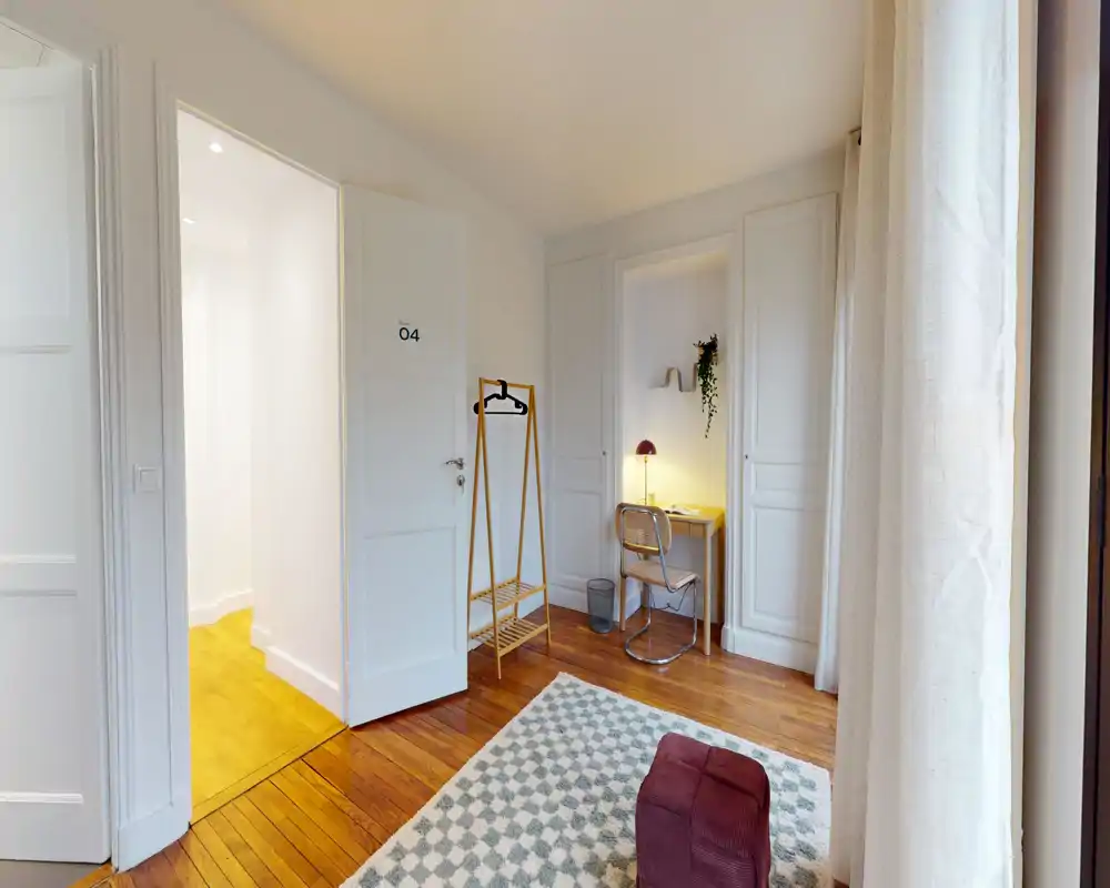 Espace de vie lumineux et soigné avec un sol en bois chaleureux, un tapis à motif et un pouf; un petit bureau avec lampe et un portant complètent l’agencement accueillant.
