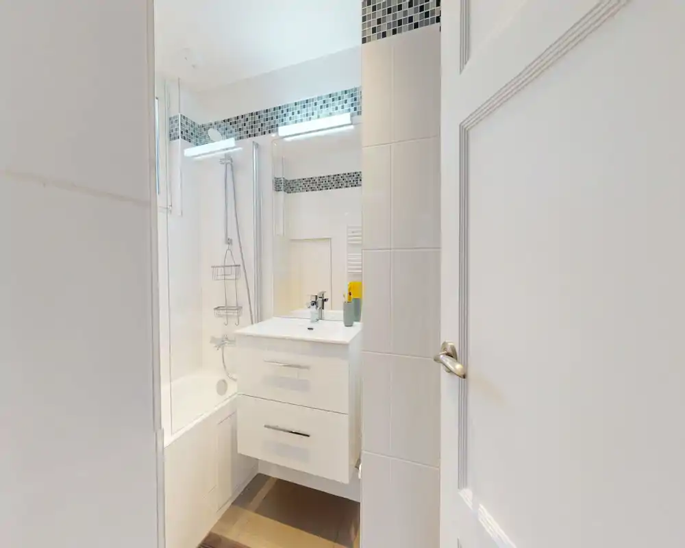 Salle de bain moderne et lumineuse avec baignoire-douche, meuble-lavabo suspendu et grand miroir encadré d’une frise en mosaïque.