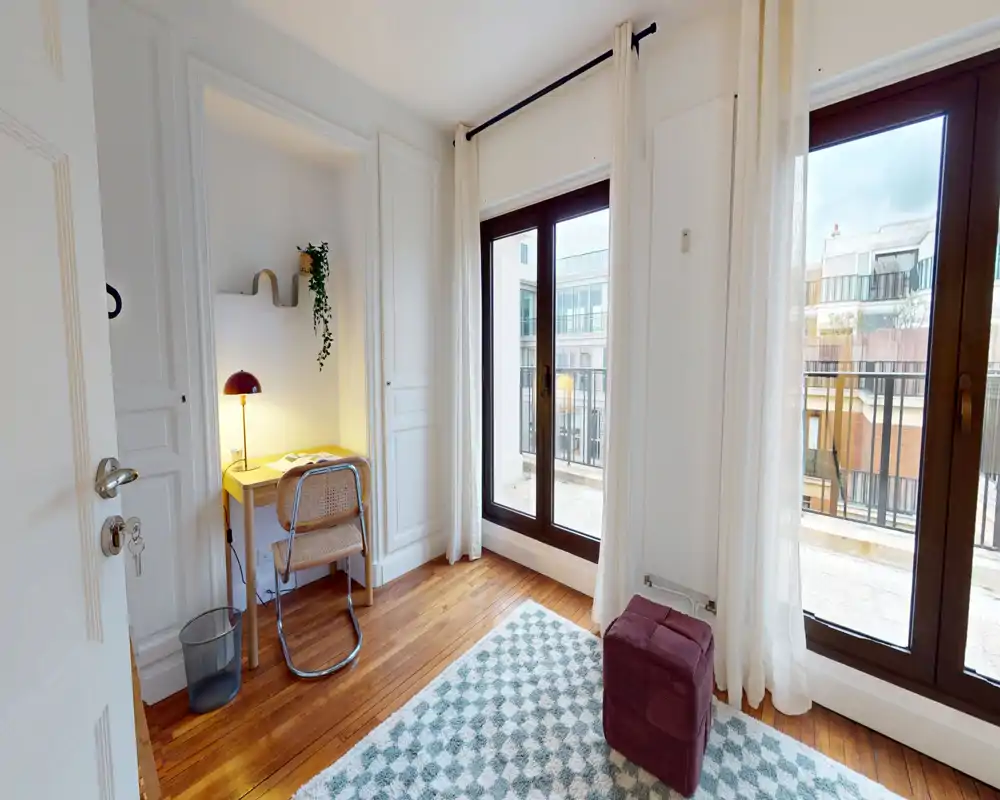 Coin de chambre lumineux et aéré avec de grandes fenêtres, un petit bureau et un pouf confortable sur un tapis à motifs. L’espace paraît moderne et apaisant grâce aux murs clairs et au parquet.
