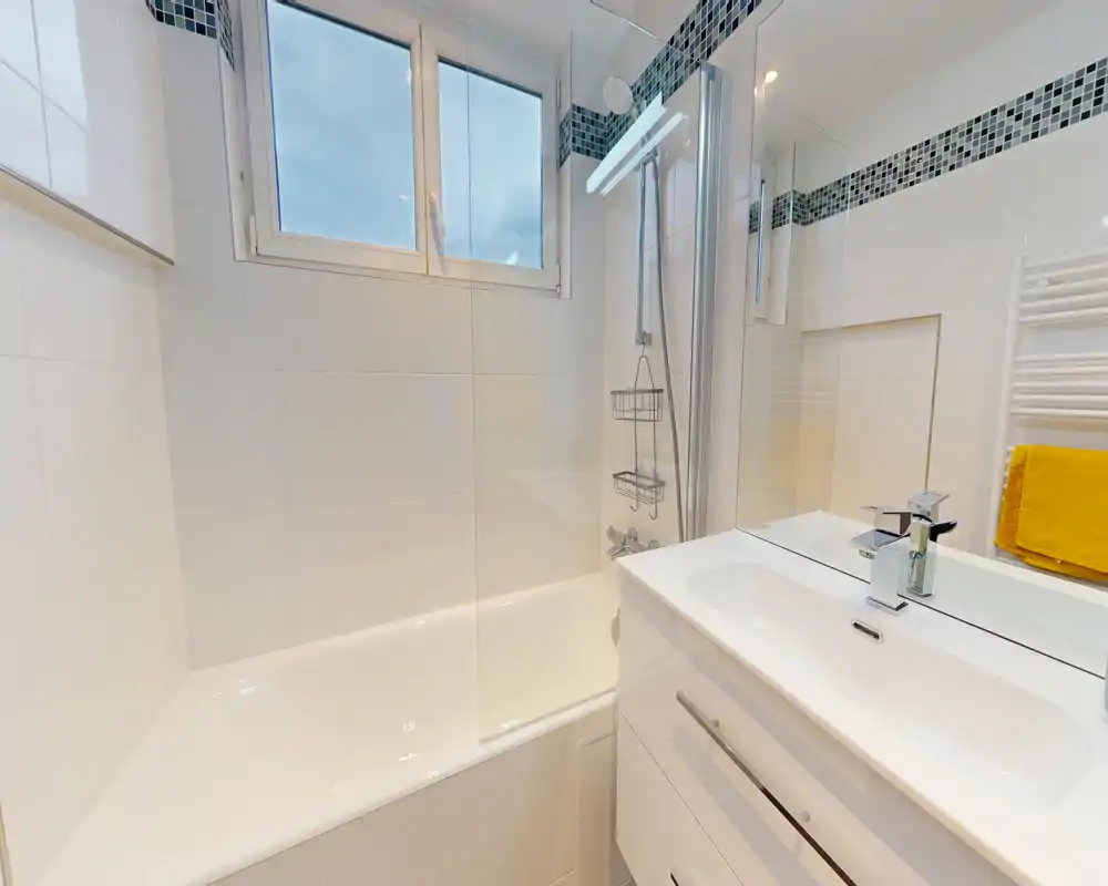Salle de bains moderne et lumineuse avec baignoire, douche et meuble vasque blanc intégré ; carrelage blanc et bandeau mosaïque pour un rendu propre et contemporain.