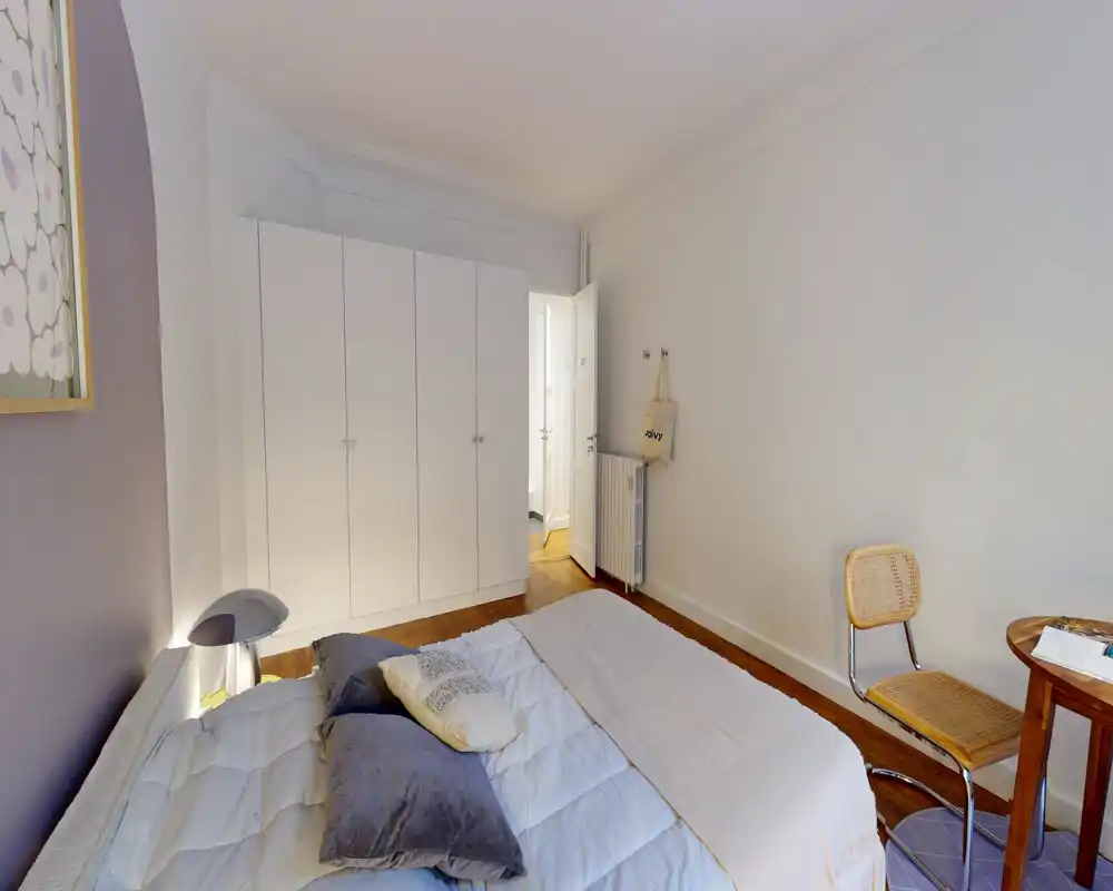 Chambre lumineuse et accueillante avec un lit double bien fait, coussins moelleux et parquet chaleureux ; placard épuré et petit bureau avec chaise dans le coin.