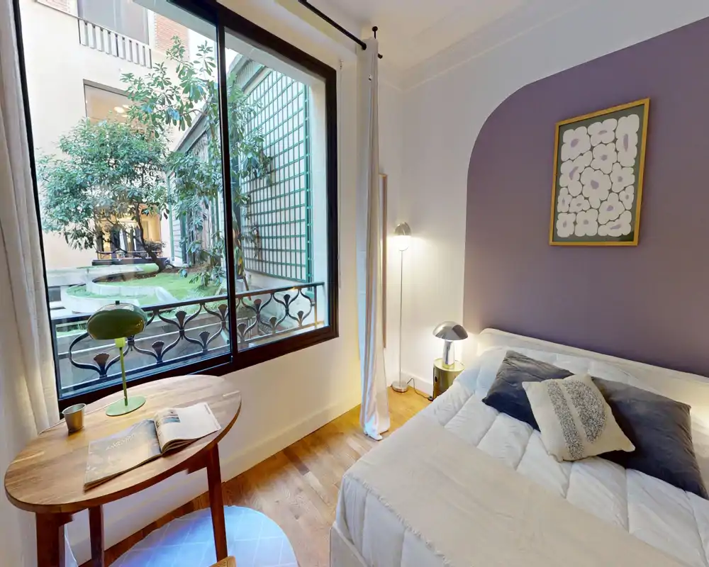 Chambre lumineuse et élégante avec un grand lit confortable, coussins décoratifs et une large fenêtre donnant sur une cour plantée. Parquet, lampe de chevet et petite table apportent une atmosphère chaleureuse.