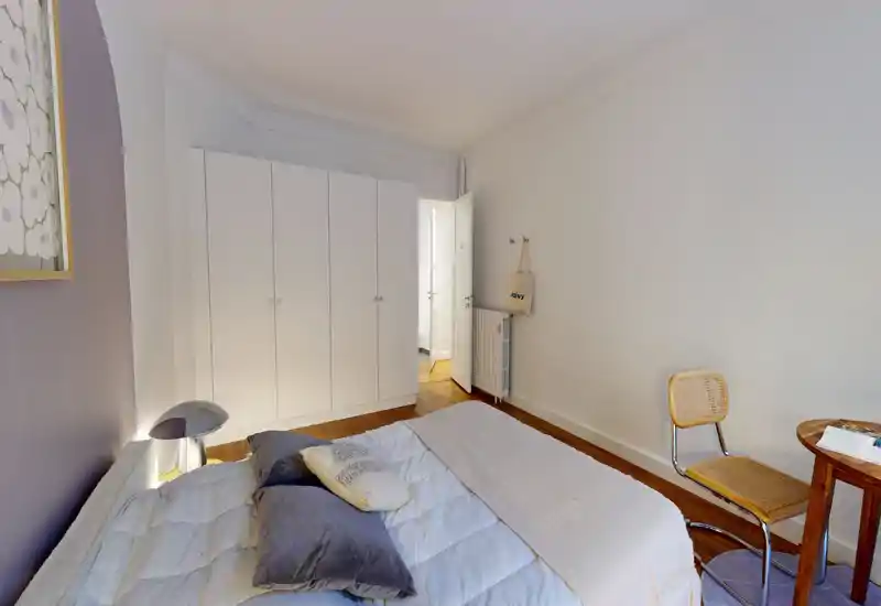 Chambre lumineuse et accueillante avec un lit double bien fait, coussins moelleux et parquet chaleureux ; placard épuré et petit bureau avec chaise dans le coin.