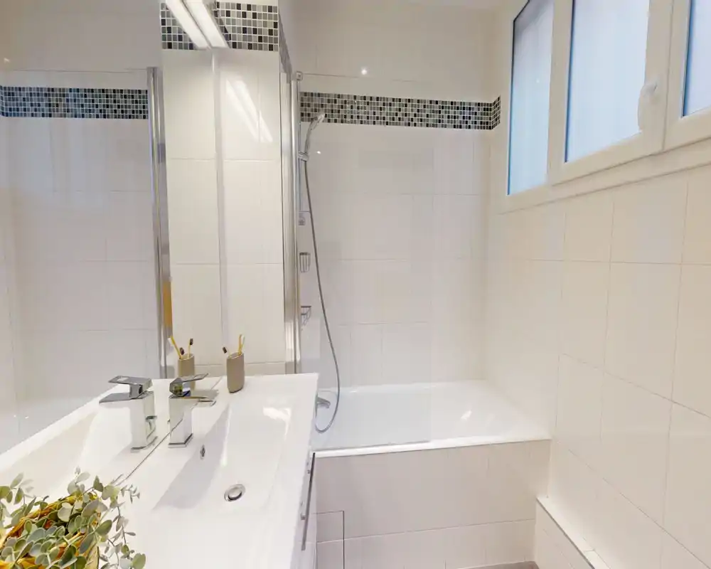 Salle de bain moderne et lumineuse avec lavabo blanc, plan de toilette et baignoire avec douchette. Carrelage propre, fenêtre dépoli pour l'intimité et grand miroir offrent une atmosphère fraîche et aérée.
