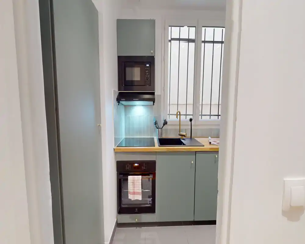 Cuisine moderne et compacte avec meubles verts, plan de travail en bois, four et micro-ondes encastrés et évier sous la fenêtre offrant une lumière naturelle.