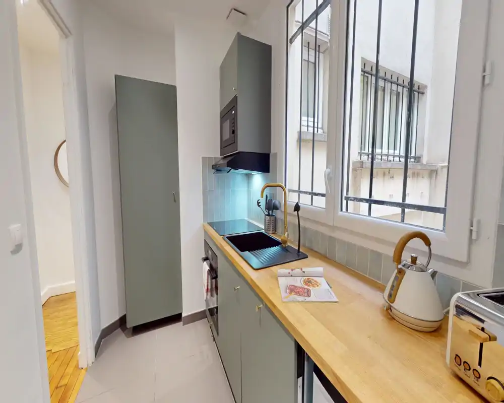 Cuisine moderne compacte avec plan de travail en bois, plaque intégrée, évier sous une grande fenêtre et rangements assortis — lumineuse et bien agencée pour un appartement urbain.