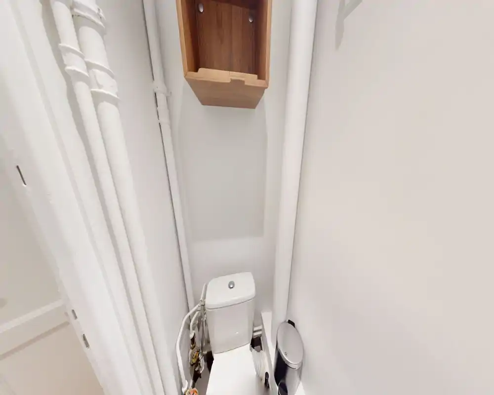 Petite salle d'eau avec WC moderne, tuyauterie apparente et petite étagère murale. Murs blancs et bonne utilisation de l'espace vertical.